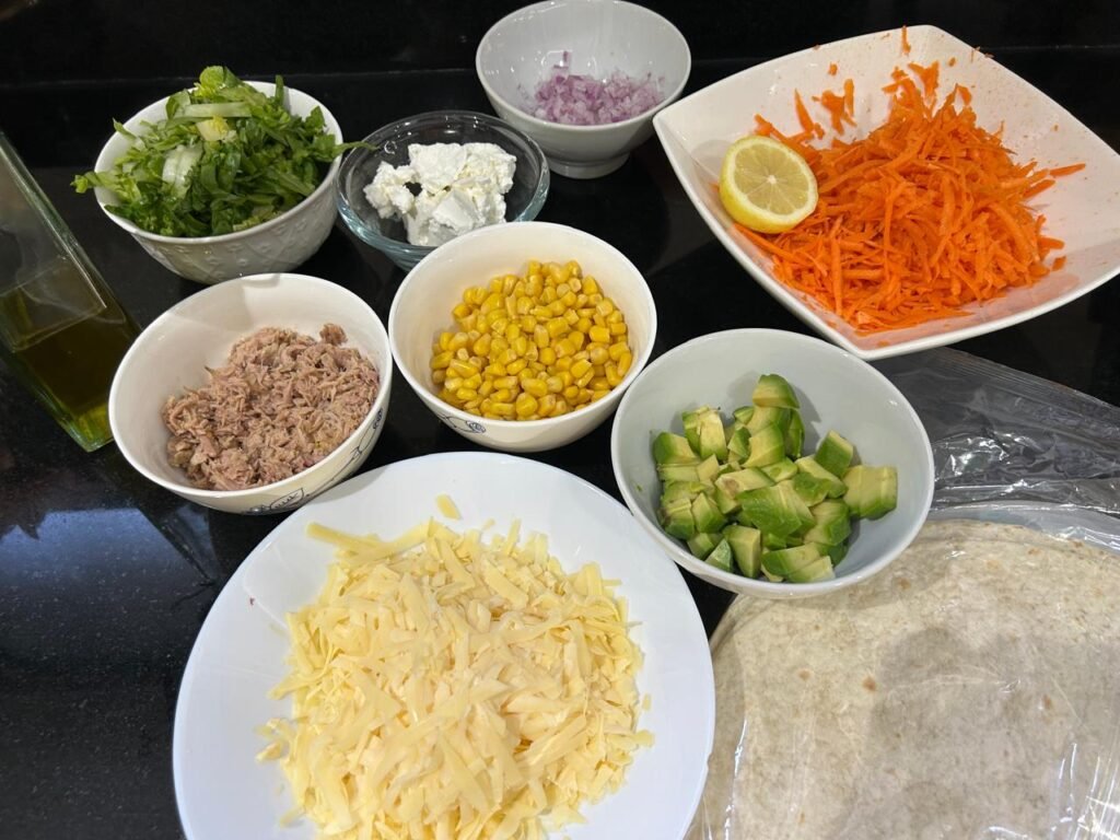 Ingredients for a tuna wrap: canned tuna, corn, avocado, grated cheese, carrot, lettuce and tortillas