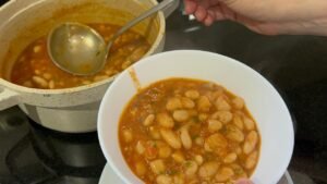 Hand ladling finished easy white bean stew from pot into white serving bowl with spoon