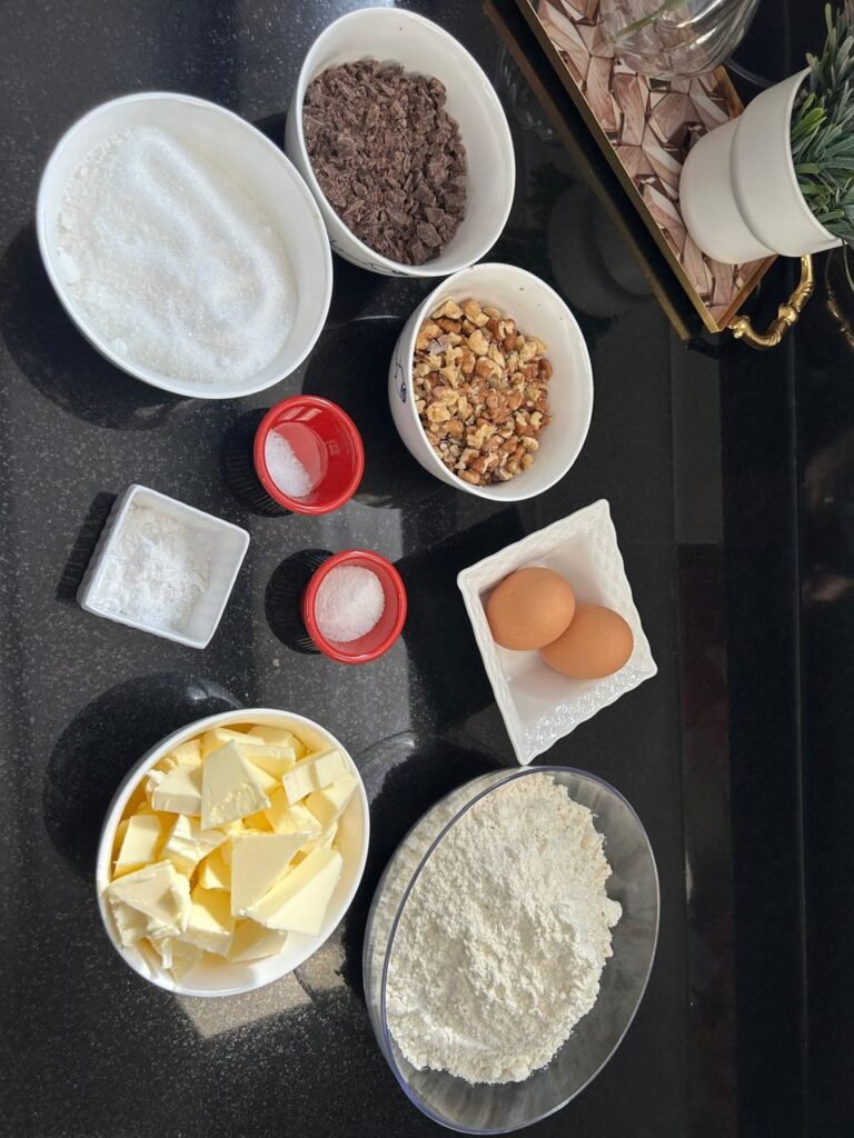 All ingredients for chocolate walnut sablé cookies arranged on dark surface: butter, sugar, eggs, flour, chocolate, walnuts