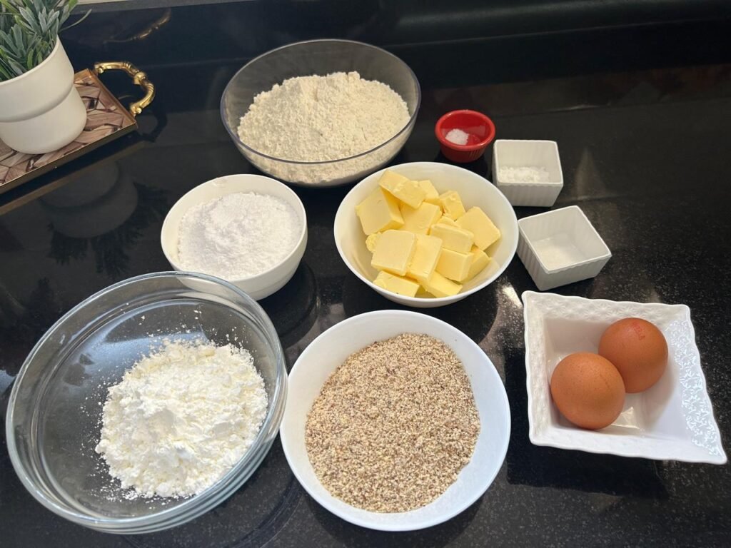 All ingredients for golden almond sablé cookies arranged on black surface: butter, eggs, flour, almonds, sugar